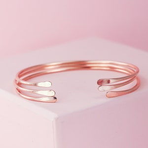 May include: A rose gold bangle bracelet with three thin bands. The bracelet is open and has a simple, minimalist design.