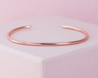 May include: A simple, thin rose gold bangle bracelet. The bracelet is made of a single, smooth piece of metal and has a polished finish.
