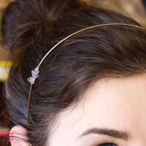 Puede incluir: Una diadema dorada con un grupo de cristales transparentes en el lateral. La diadema es fina y delicada, y la lleva una persona con el pelo castaño.