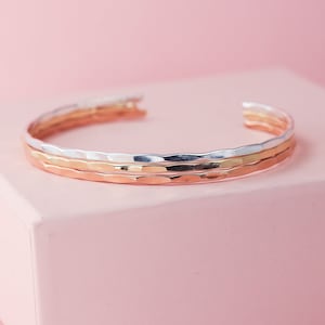 May include: Three stacked hammered metal bangles, one silver, one gold, and one rose gold. The bangles are on a pink surface.