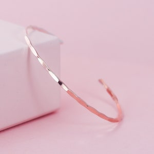 May include: A delicate rose gold hammered bangle bracelet. The bracelet is thin and has a simple, minimalist design.