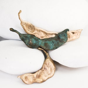 May include: Three abstract sculptures made of metal, resembling pods or seed casings. The sculptures are in shades of gold and green, and are arranged on a white surface.
