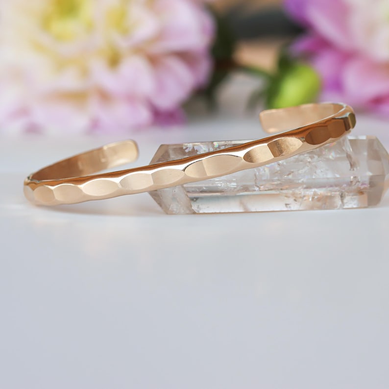 May include: A gold hammered cuff bracelet. The bracelet is made of a smooth, shiny metal and has a hammered texture. The bracelet is open ended and can be adjusted to fit most wrists.