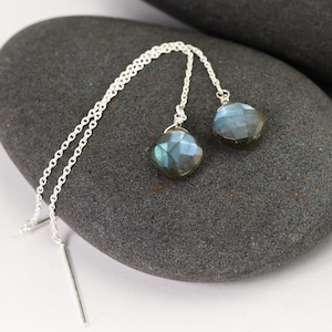 May include: A pair of silver threader earrings with faceted, iridescent labradorite gemstone drops. The earrings are displayed on a dark gray stone, showcasing the blue and green hues of the gemstones.