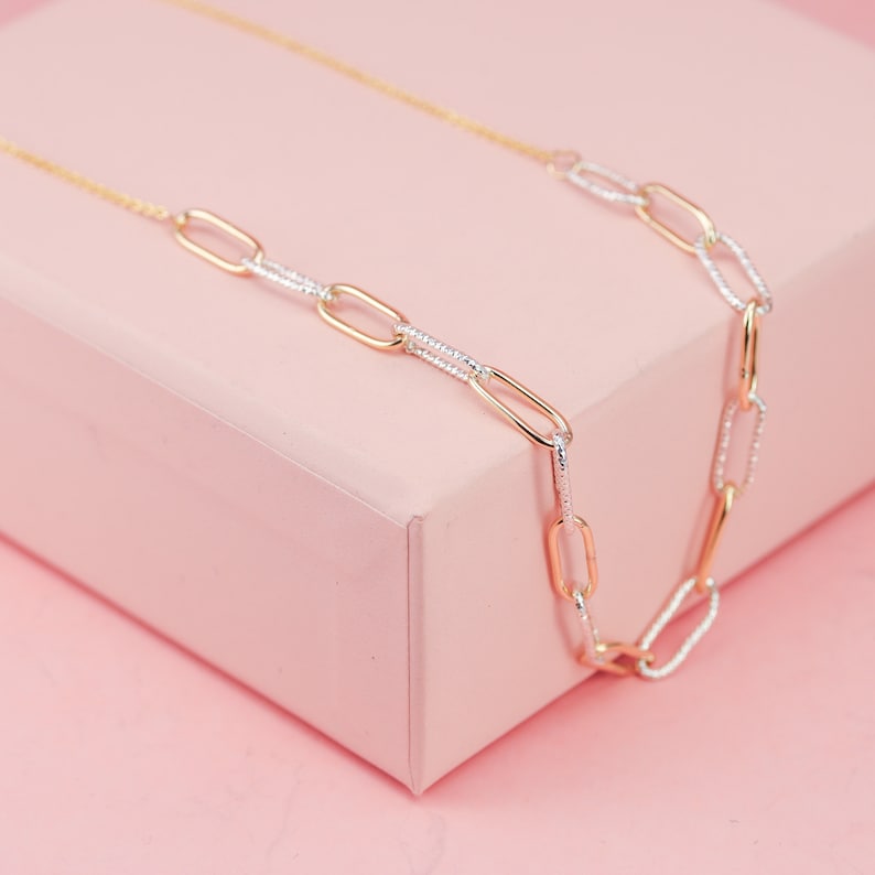 May include: A gold and silver chain necklace with oval links. The chain is on a pink surface.