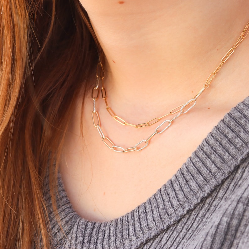 May include: A layered gold chain necklace with a delicate, textured design. The necklace is made of two chains, one with a slightly larger link than the other. The necklace is worn by a person with long brown hair.