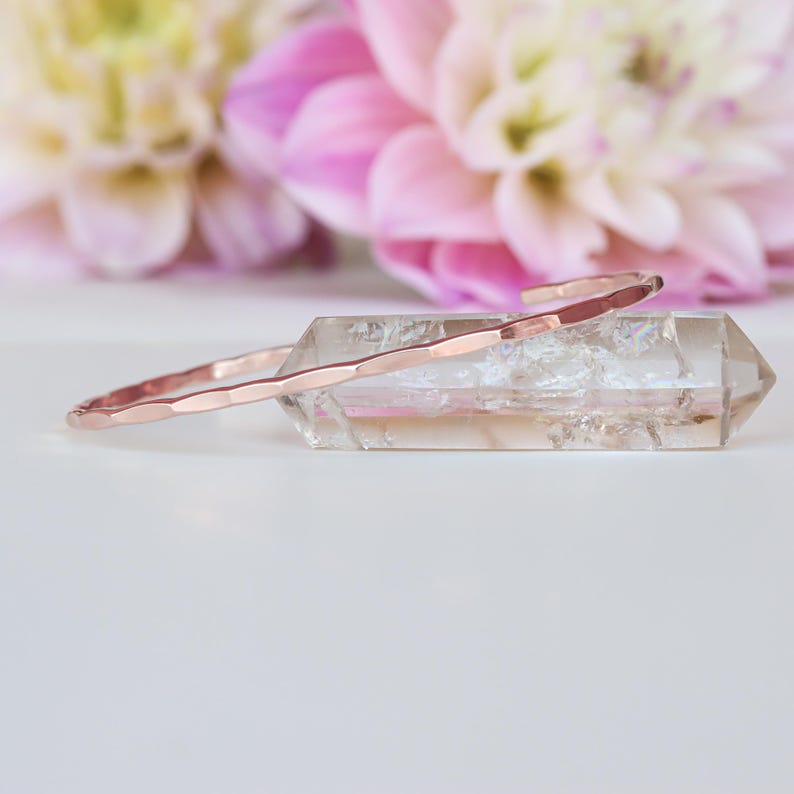 Pode incluir: Um bracelete de ouro rosa martelado descansando sobre um cristal de quartzo transparente. O bracelete &eacute; fino e delicado, com um design simples e elegante.