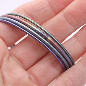 May include: Close-up of several thin, iridescent metal bangles in shades of purple, green, and silver. The bangles are stacked together, creating a layered effect. The image is well-lit, highlighting the metallic sheen.