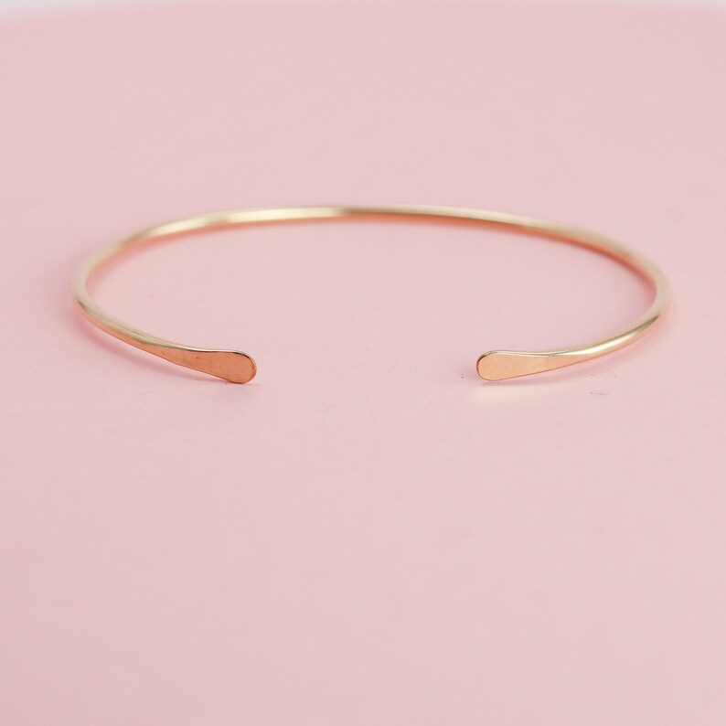 May include: A simple gold bangle bracelet with rounded ends. The bracelet is made of a thin, smooth metal and has a minimalist design.
