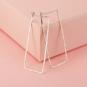 May include: A pair of silver rectangular hoop earrings. The earrings have a hammered texture and a simple, modern design. They are displayed on a pink surface against a matching pink background. The earrings are lightweight and suitable for everyday wear.