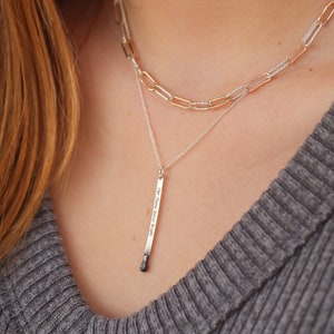 May include: A silver necklace with a silver matchstick pendant that reads "One flame burns so bright". The necklace is layered over a gold chain necklace.
