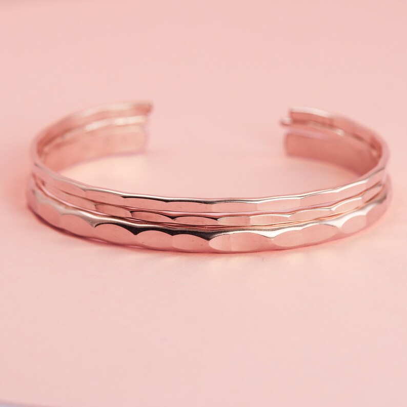 Pode incluir: Tr&ecirc;s pulseiras de metal martelado em ouro rosa empilhadas. As pulseiras s&atilde;o lisas e t&ecirc;m uma forma ligeiramente arredondada.