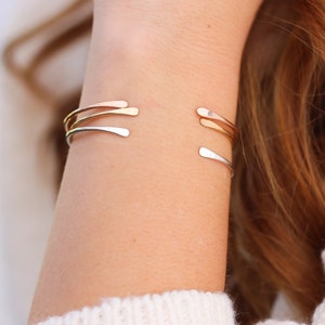 May include: Three gold bangle bracelets stacked on a person's wrist. The bracelets are thin and have a simple, modern design.