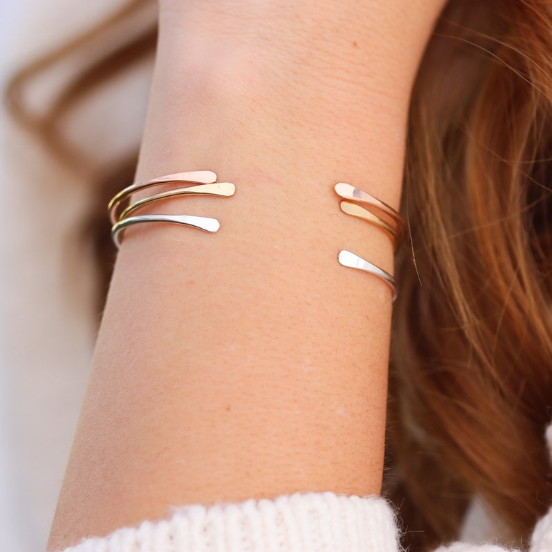 May include: Three gold bangle bracelets stacked on a person's wrist. The bracelets are thin and have a simple, modern design.