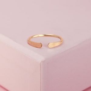 May include: A simple gold ring with a slightly rounded, open design. The ring is shown on a pink surface.