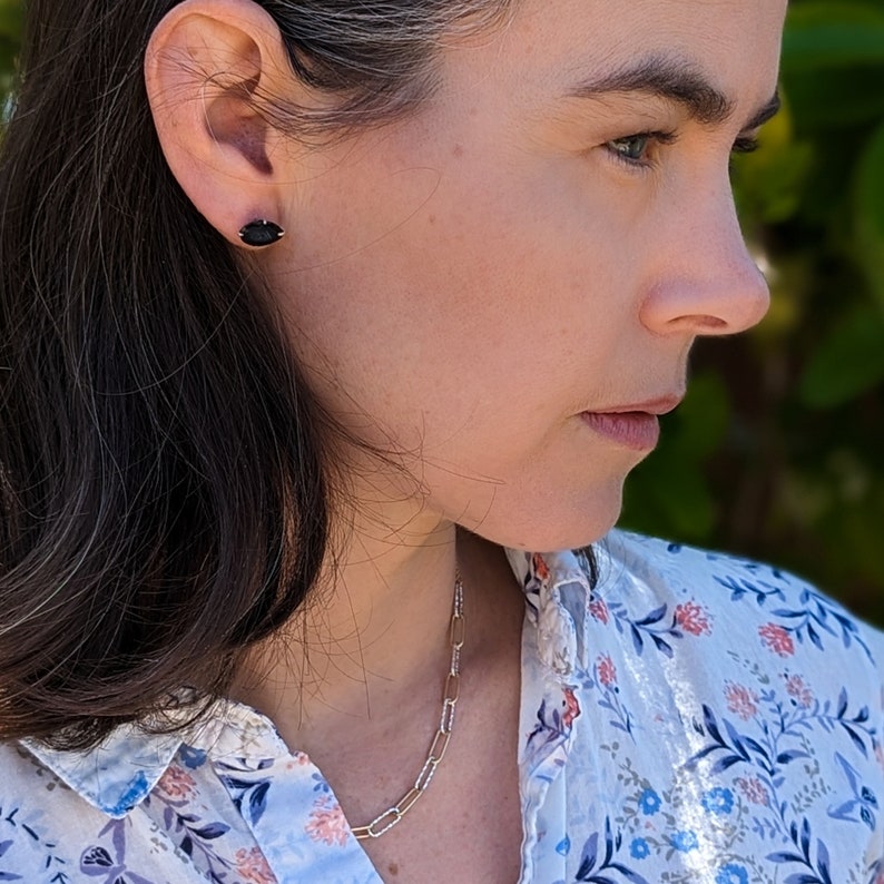 May include: A woman with dark hair wearing a white blouse with a floral pattern and a silver chain necklace. She is wearing a small black oval earring in her left ear.