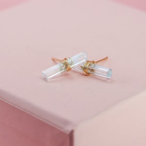 May include: Stud earrings with clear, rectangular crystals, wrapped with gold-coloured wire. The earrings are set against a soft pink background, creating a delicate aesthetic. The crystals appear to be approximately 1.3 cm in length.