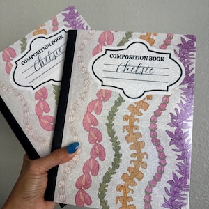 May include: Two composition notebooks with floral designs. The covers feature a white background with pink, green, orange, and purple floral patterns. Each notebook has the text "COMPOSITION BOOK" and the name "Chelsie" on the cover.