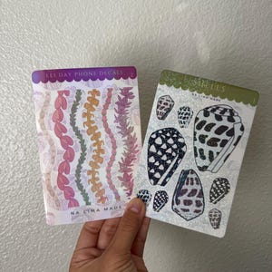 May include: Two sticker sheets are held in a hand. One sheet has a purple header that reads "Lei Day Phone Decals" and features pink, orange, and purple floral designs. The other sheet has a green header and features seashell designs.