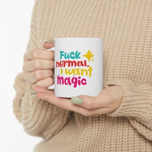 Könnte beinhalten: Weiße Keramik-Tasse mit dem Text "Fuck normal, I want magic" in farbenfrohen, verspielten Buchstaben. Eine gelbe Stern-Grafik befindet sich neben dem Text. Die Tasse wird von einer Person gehalten, die einen beigen Pullover trägt.