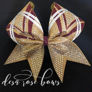 May include: A gold and maroon cheer bow with white stripes and rhinestones. The bow is tied with a black elastic band.