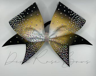 May include: A black, gold, and white cheer bow with iridescent rhinestones. The bow has a ombre design with black at the edges and gold in the center.