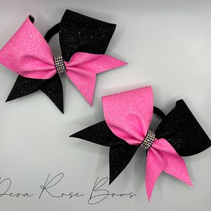 May include: Two cheer bows with black and pink glitter fabric. The bows have a rhinestone center and are tied with black elastic.