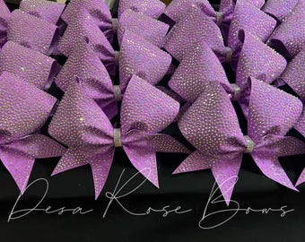 May include: A group of purple cheer bows with iridescent rhinestones. The bows are arranged in rows on a black surface. The text "Desa Rose Bows" is visible at the bottom of the image.