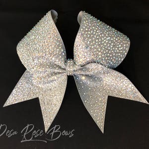 May include: A large silver cheer bow with iridescent rhinestones. The bow is made of glitter fabric and has a pointed shape.