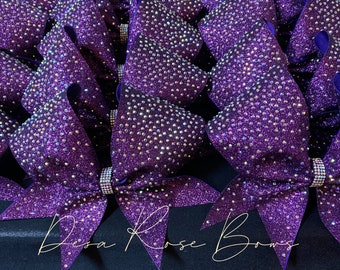 May include: A close-up of multiple purple cheer bows with iridescent rhinestones. The bows are arranged in a pile, with the text "Dosa Rose Bows" visible at the bottom of the image.
