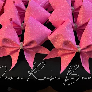 May include: A group of pink glitter cheer bows with silver rhinestone accents. The bows are arranged in a row and are all the same size. The bows are on a black surface with the text "Desa Rose Bows" written in white.
