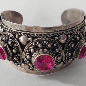 Puede incluir: Un brazalete de plata con intrincados diseños florales y tres piedras preciosas rosas.