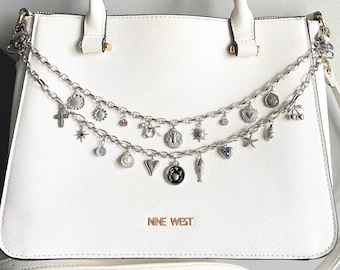 May include: White leather handbag with a silver chain strap adorned with multiple charms, including a heart, a fish, a star, and a sun. The bag has the words "Nine West" printed on the front.
