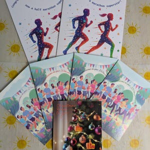 Op de afbeelding: Een verzameling wenskaarten. Sommige kaarten tonen hardlopers met sterpatronen en de tekst "Gefeliciteerd, je bent nu een halve marathon superster!" en "Gefeliciteerd, je bent nu een marathon superster!". Andere kaarten tonen hardlopers en de tekst "Gefeliciteerd met je 25e deel". Een kerstkaart met een versierde boom is ook inbegrepen.