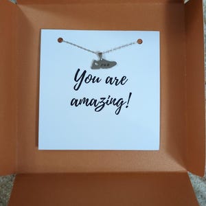 May include: A white card with the text "You are amazing!" and a silver necklace with a shoe pendant that says "run" on it. The card and necklace are in a brown box.