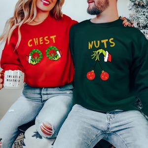 May include: A couple wearing matching Christmas sweaters. The woman's sweater is red and says "CHEST" with two green wreaths and green hands holding the wreaths. The man's sweater is green and says "NUTS" with a green hand holding two red ornaments.