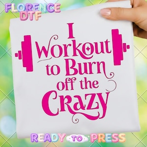 May include: White t-shirt with the text "I Workout to Burn off the Crazy" in bright pink. The design includes barbell graphics on either side of the text. The words "Florence DTF" and "Ready to Press" are also visible.