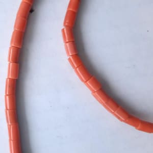 May include: Close-up of a necklace made of coral-colored, cylindrical beads. The beads are strung together, forming a curved line against a white background. The coral beads have a smooth, polished appearance.