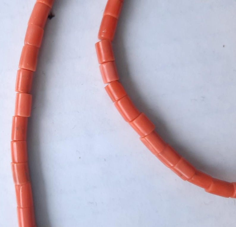May include: Close-up of a necklace made of coral-colored, cylindrical beads. The beads are strung together, forming a curved line against a white background. The coral beads have a smooth, polished appearance.