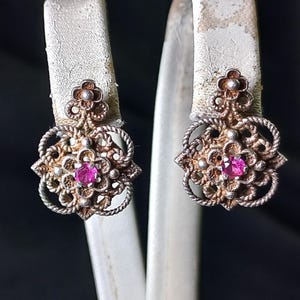 Peut inclure: Une paire de boucles d'oreilles vintage en argent avec un motif floral. Chaque boucle d'oreille présente une pierre précieuse rose centrale, entourée de filigranes complexes et de détails en forme de corde. Les boucles d'oreilles sont présentées sur une surface blanche.