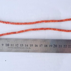 May include: A necklace made of small, rectangular, coral-colored beads. The necklace is laid out on a white surface next to a metal ruler. The ruler measures up to 32 inches.