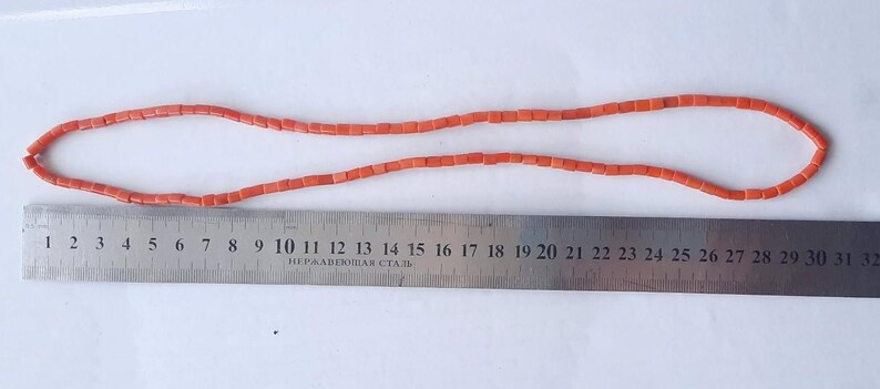 May include: A necklace made of small, rectangular, coral-colored beads. The necklace is laid out on a white surface next to a metal ruler. The ruler measures up to 32 inches.