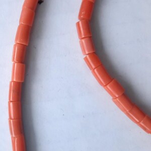 May include: Close-up of a coral necklace. The necklace features cylindrical beads in a vibrant, reddish-orange hue. The beads are strung together, creating a curved line against a white background. The coral beads are evenly sized and shaped.