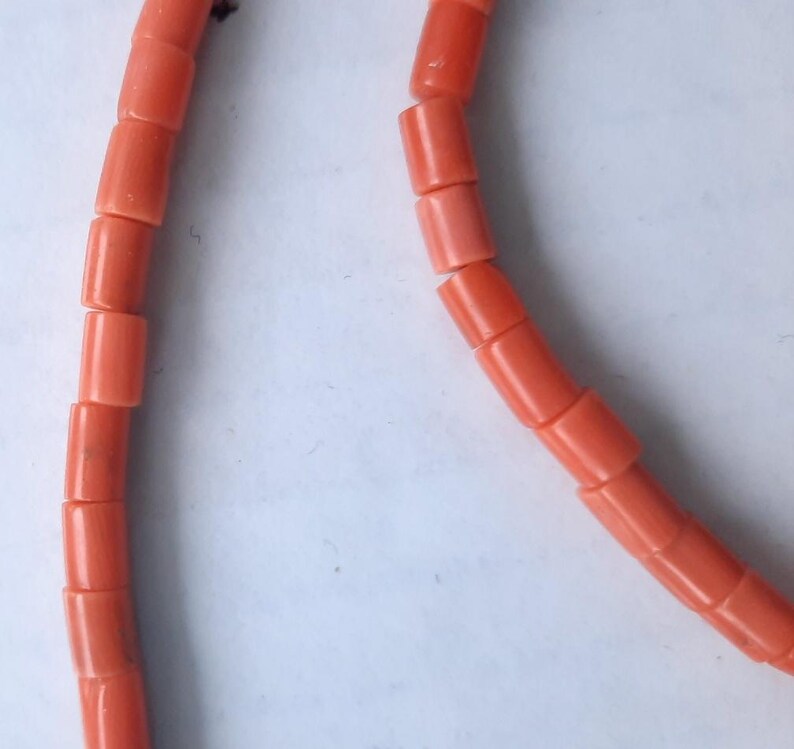 May include: Close-up of a coral necklace. The necklace features cylindrical beads in a vibrant, reddish-orange hue. The beads are strung together, creating a curved line against a white background. The coral beads are evenly sized and shaped.