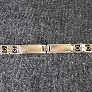 May include: A gold-coloured bracelet with a geometric design. The bracelet features rectangular and diamond-shaped links, connected by smaller, decorative elements. The clasp is visible at the end. The bracelet is displayed on a dark grey surface.