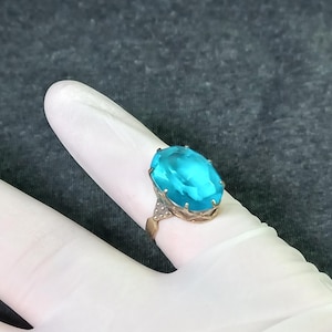 May include: A gold-toned ring featuring a large, oval-cut, bright blue gemstone. The ring's band has a decorative design with small, clear accent stones. The ring is displayed on a white surface against a gray background.