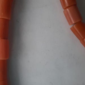 May include: Close-up of a strand of coral beads. The beads are cylindrical, with a smooth, polished surface. The color is a warm, salmon-orange. The beads are strung together, forming a necklace or bracelet.