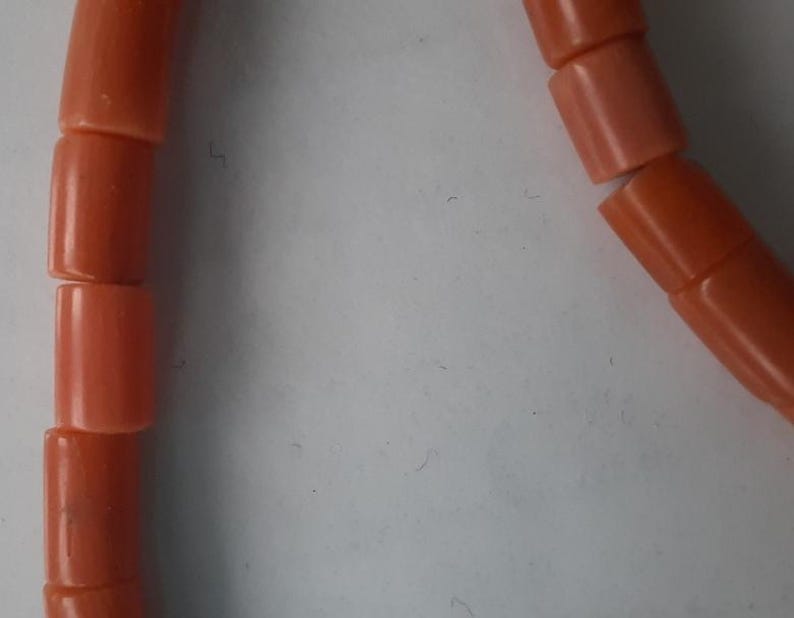 May include: Close-up of a strand of coral beads. The beads are cylindrical, with a smooth, polished surface. The color is a warm, salmon-orange. The beads are strung together, forming a necklace or bracelet.
