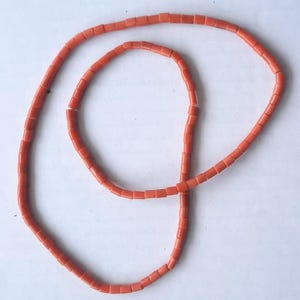May include: A necklace composed of small, rectangular coral beads in a vibrant orange hue. The beads are strung together, forming a continuous loop. The necklace is displayed against a plain white background.
