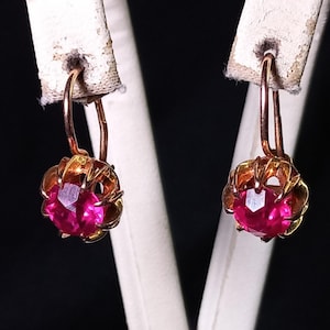 Puede incluir: Un par de pendientes vintage con una gema rosa brillante engastada en un diseño de filigrana dorada. Los pendientes tienen cierre de palanca. Fondo oscuro.